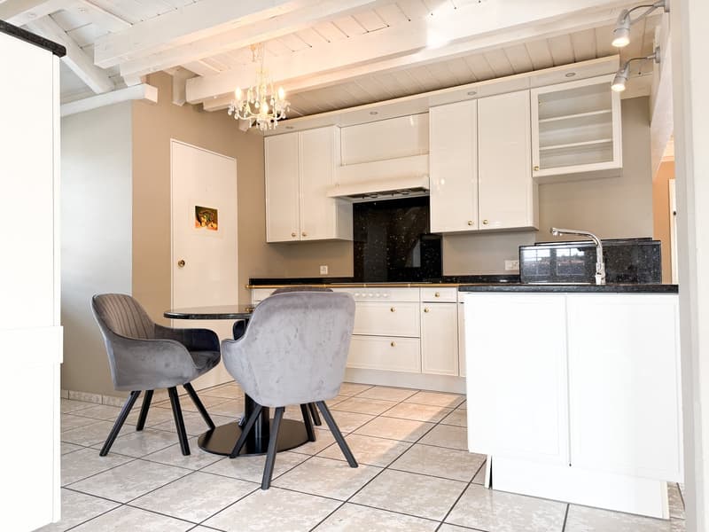 Modern, compact kitchen with white cabinets, marble countertop, stainless steel sink, chandelier, tiled floor, small round table, two chairs, and a black microwave on the counter.