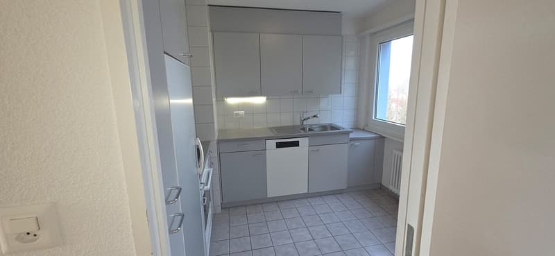 A modern kitchen with white cabinets, countertops, a stainless steel sink with a faucet, a dishwasher, an electric oven, and white tiled flooring.