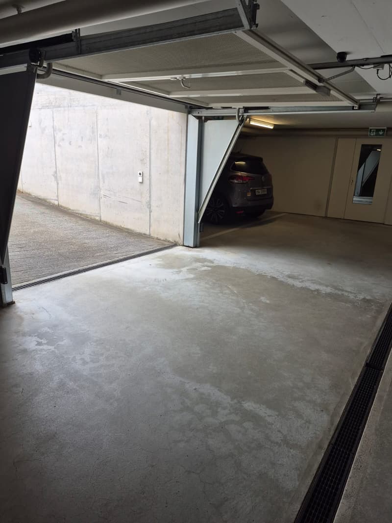 The image shows the interior of a garage with a concrete floor and a car parked inside. The garage has an overhead door and appears to be a part of a larger building or structure.
