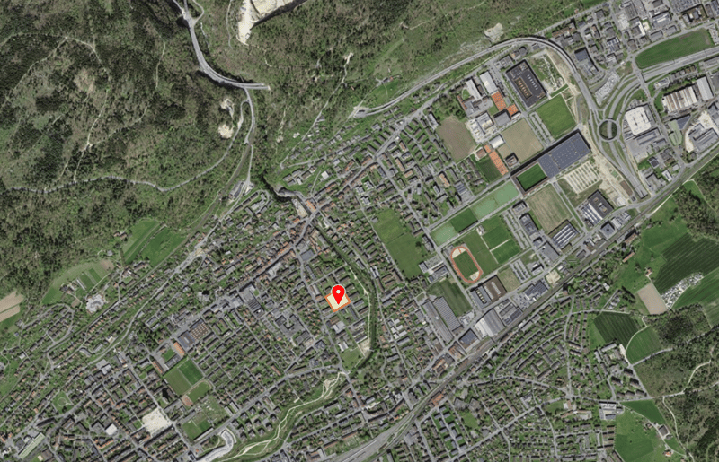Aerial view of a town with many buildings, roads, and green spaces. The town is surrounded by trees and mountains.