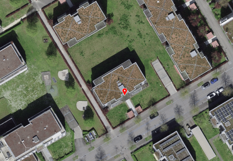 Aerial view of a residential area with multiple buildings, green lawns, and a central pathway. Several cars are parked along the road.