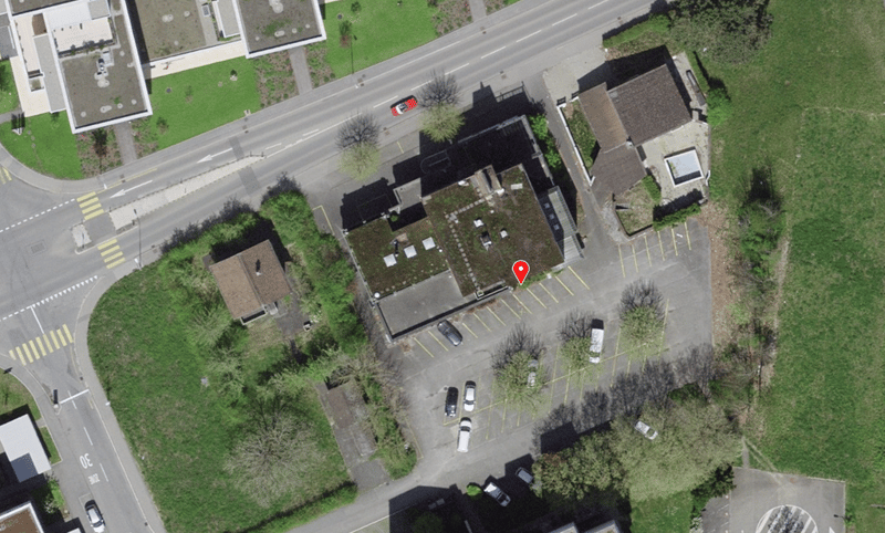 Aerial view of a parking lot with several cars parked, a building with a green roof, and a red marker indicating a specific location.