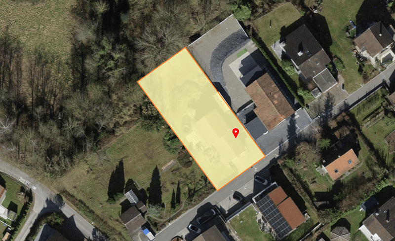 Plot of land with a rectangular shape, surrounded by houses and greenery, with a driveway leading to the plot.
