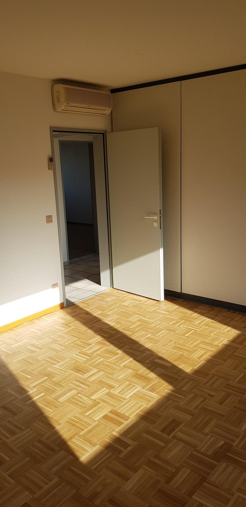 empty room, wooden parquet flooring, white walls, open door, large window, air conditioning unit