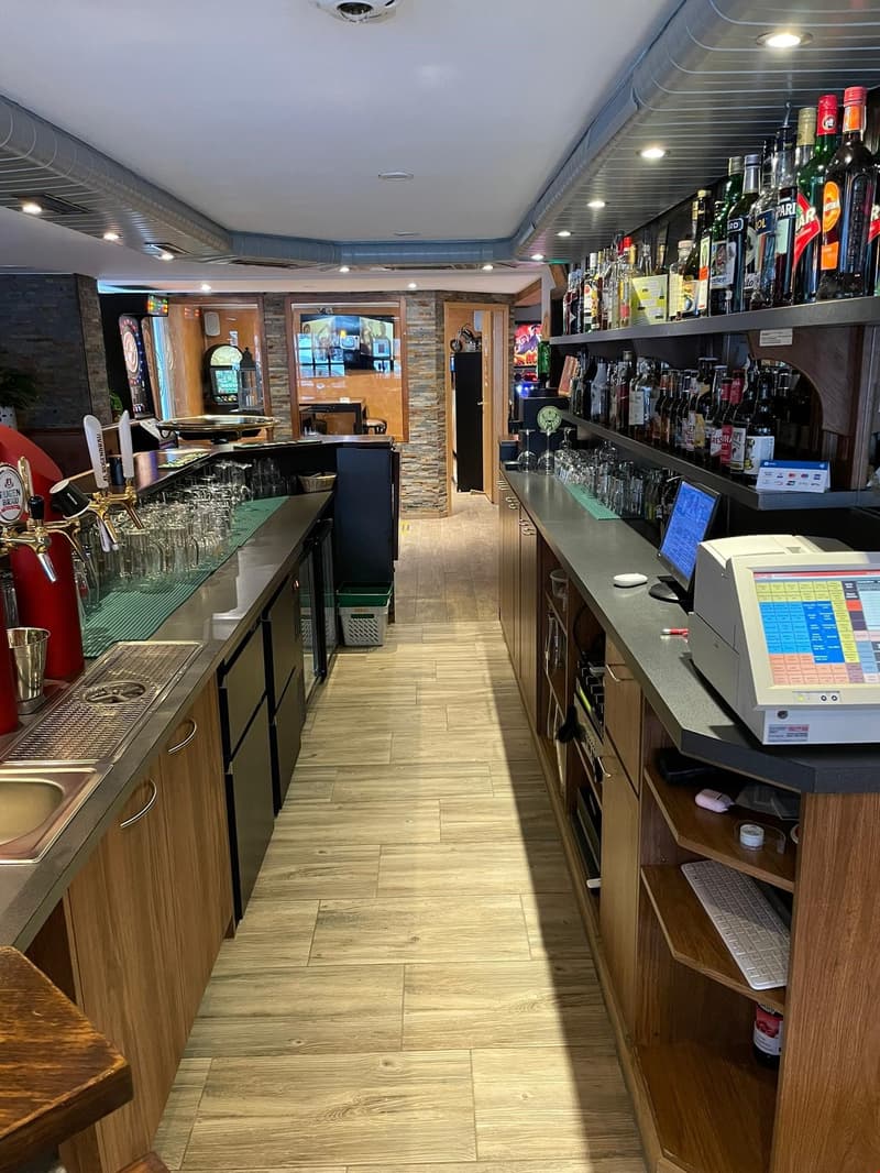 Wooden counter with glasses, bottles, dartboard, and kitchen equipment, shelves with liquor bottles, cash register, and computer.