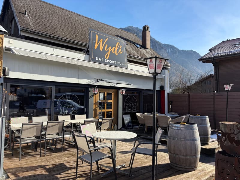 Exterior of a building with outdoor seating area and wooden deck, surrounded by tables and chairs, barrels, outdoor lamps, wooden signboard reading 'Wydi', and mountains in the background.