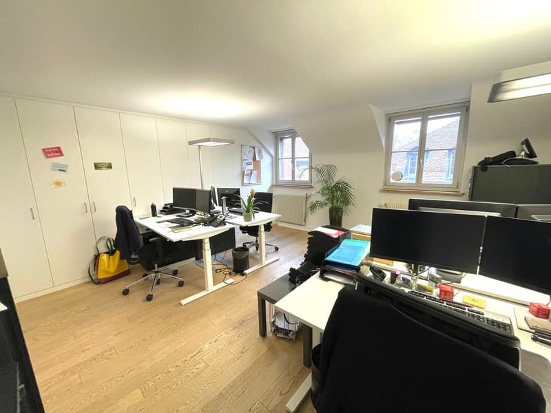 The image shows a spacious office space with multiple workstations. The room has hardwood floors, white walls, and large windows that provide natural light. There are several desks with computers, office chairs, and other office equipment such as printers