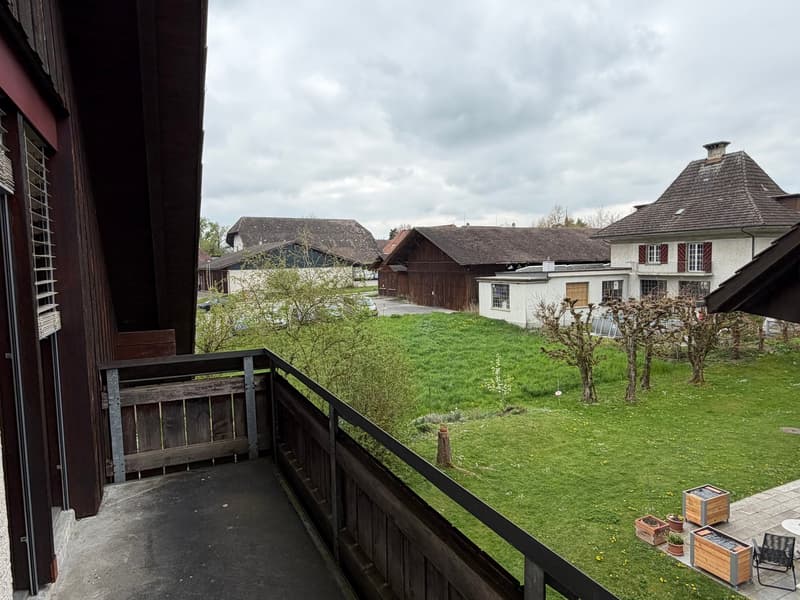 Balcony with wooden railing, view of a grassy field with trees and houses in the background