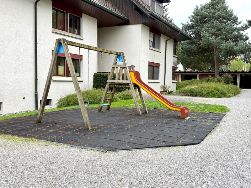Playground with swings and slide, gravel ground, white building in the background