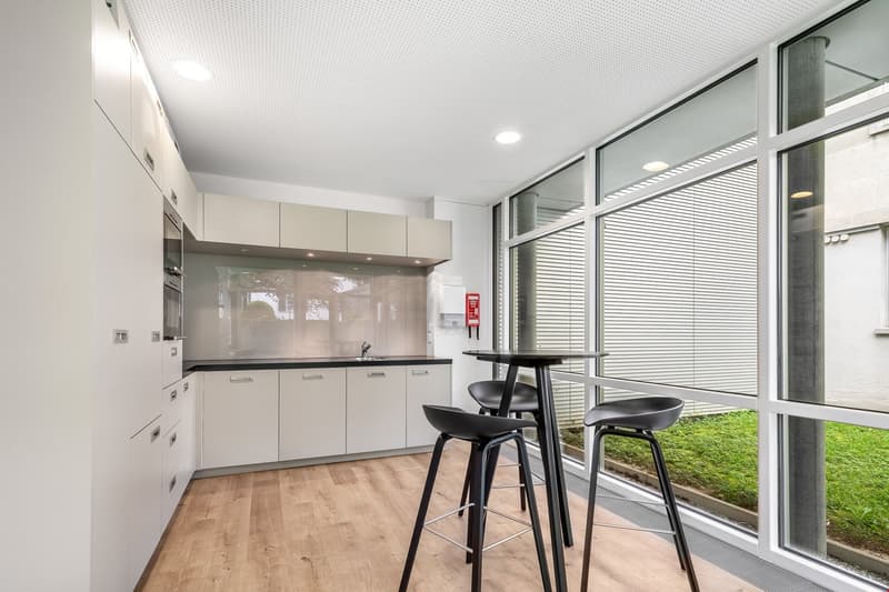 Modern kitchen with white cabinets, black countertops, stainless steel appliances, and a dining area with a high table and stools. Large windows provide a view of the outdoor greenery.
