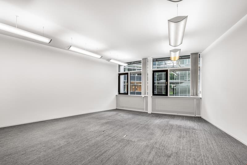 Large open office space with white walls, gray carpeted floor, and large windows providing natural light. The room is equipped with modern lighting fixtures and appears to be a commercial or office setting.