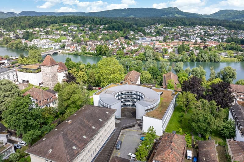 Aerial view of a picturesque city nestled in a valley surrounded by mountains and forests. The city features a mix of historic buildings, modern architecture, and lush greenery. A river runs through the center of the city, adding to the scenic landscape.