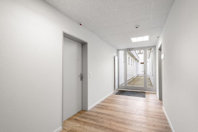 Empty hallway with white walls, wooden floor, glass doors, and a black mat at the entrance.