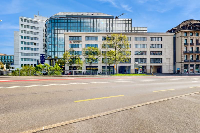 Multi-story commercial building with glass facade, surrounded by trees and located on a busy street with parking spaces
