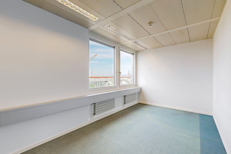 The image shows a spacious office space with large windows providing a view of the city skyline. The room has a clean, minimalist design with white walls, a suspended ceiling, and a tiled floor. There are radiators along the walls, suggesting this is a fu