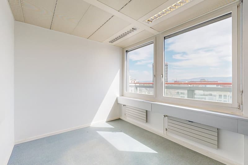 Bright and spacious office space with large windows offering a view of the city skyline. The room has a clean, minimalist design with white walls and a tiled floor. There are built-in storage units along the window sill.