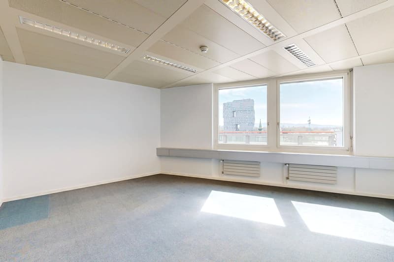 Spacious office room with large windows, white walls, suspended ceiling with recessed lighting, and built-in storage units along the walls.