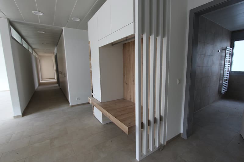 The image shows a long hallway with white walls, a tiled floor, and built-in storage units or closets along the walls. There is a wooden bench or seating area visible in the hallway, and a doorway leading to another room can be seen at the end of the hall