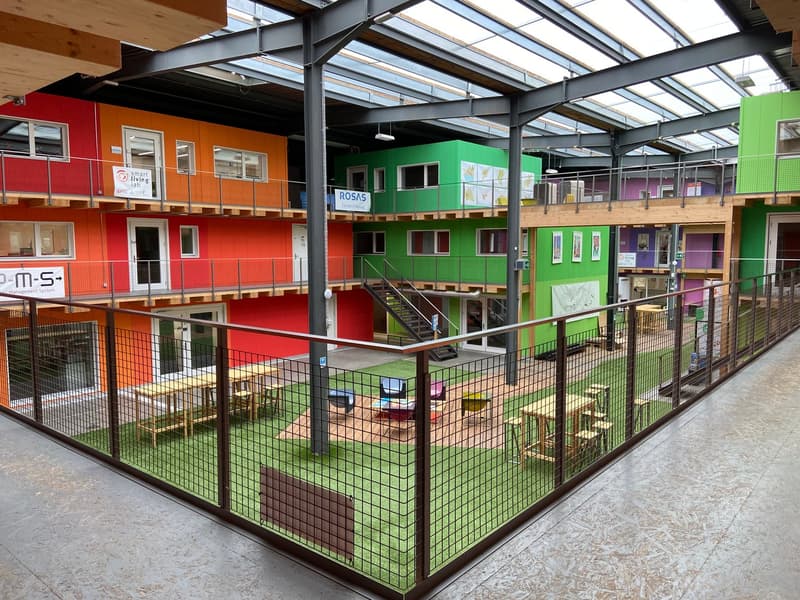 Open area with tables, chairs, and seating. Multi-colored modular buildings on the left, right, and in the background. Raised ground floor with steel beams and large windows. Glass roof.