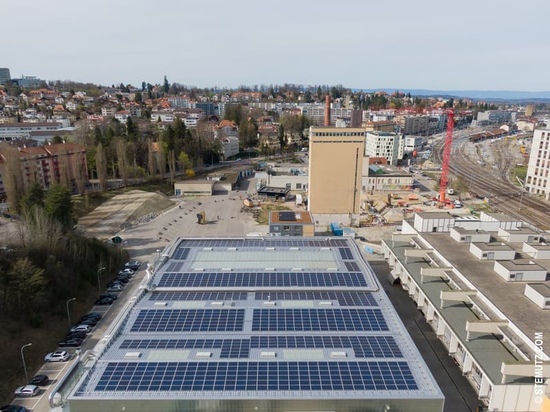 Industrial building with solar panels on the roof, multiple levels, large windows, surrounded by trees and construction site