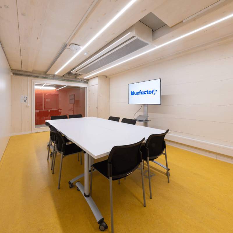 Conference room with a long white table, black chairs, and a mounted monitor displaying 'bluefactor', wooden walls and floor.