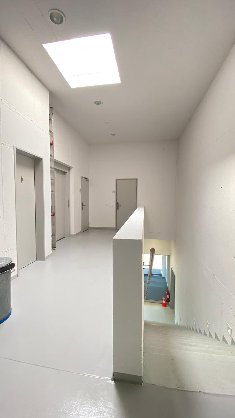 Empty hallway with white walls, white floor, two elevators, two doors, a staircase, and a trash bin.