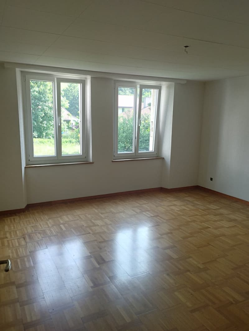 Spacious living room with large windows overlooking a garden, hardwood flooring, and white walls.