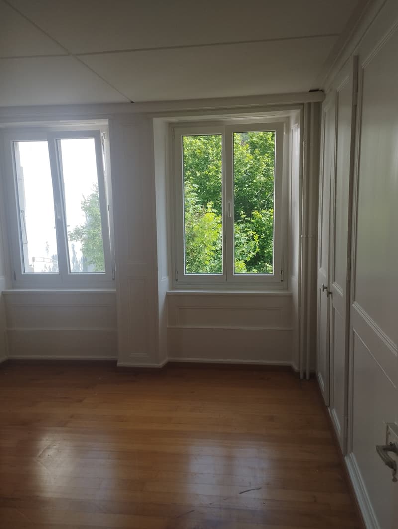 Spacious room with large windows overlooking a lush, green outdoor area. The room has hardwood floors and white walls, creating a bright and airy atmosphere.