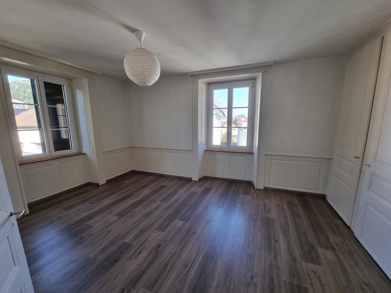 Empty room with wooden flooring, white walls, large windows, and white ceiling with hanging lamp