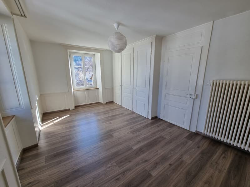 Large empty room with white walls, wooden flooring, a window, and a radiator on the right side