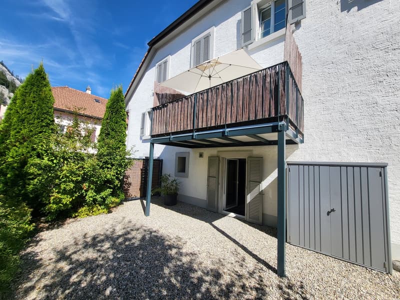 white house, balcony with wooden railing, gravel driveway, gray garage door, potted plant