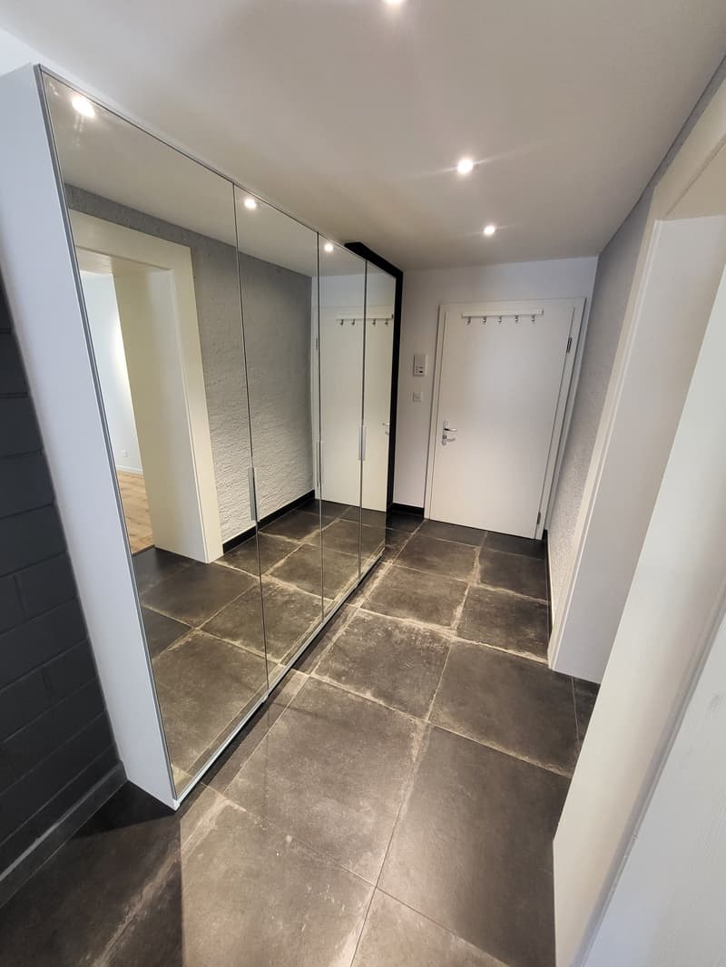 Empty hallway with large mirror, tiled floor, white walls, and a door.
