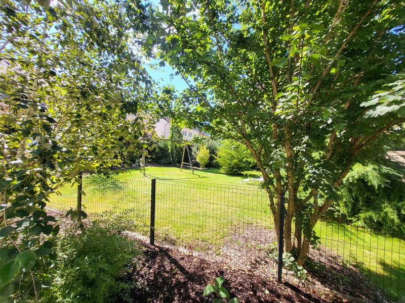 fenced garden with green grass, trees, plants, swing, and a house in the background
