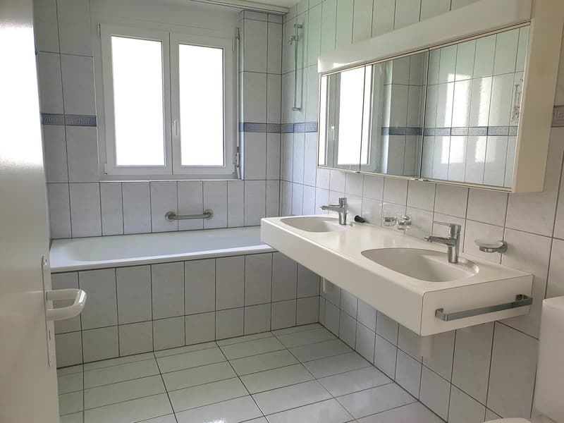 Bathroom with white tiles, bathtub, double sink, mirror, and window.