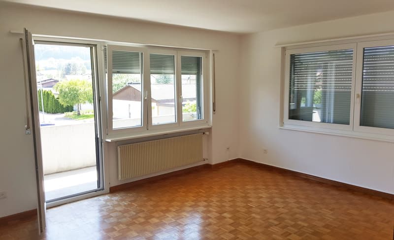 Large empty room, wooden floor, two windows with sliding doors, central heating radiator