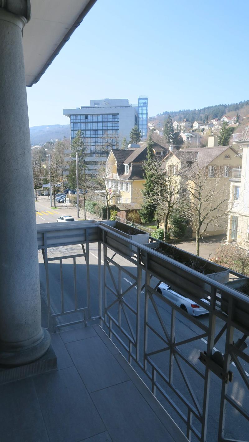 Balcony with metal railings, concrete floor, metal column, view of the street, parked cars, trees, and buildings