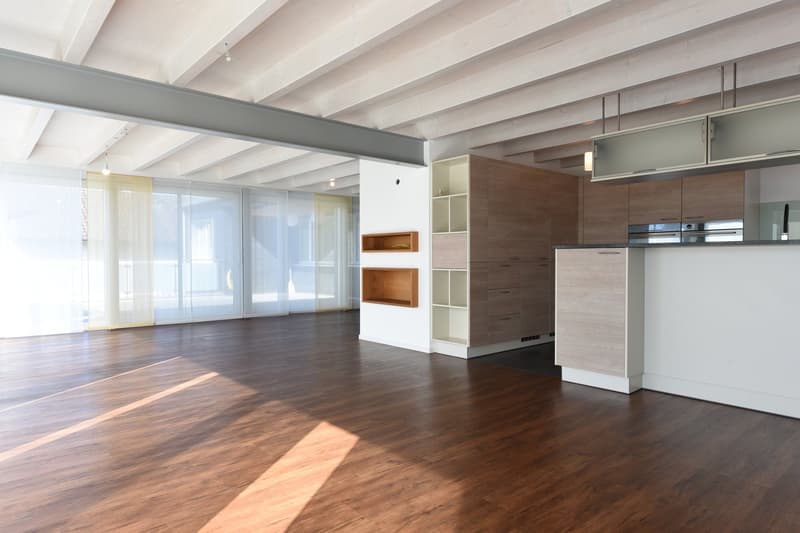 Open space with wooden floors, white walls, large windows, modern kitchen with white cabinets and countertops, wooden shelves, and a ceiling with exposed beams.