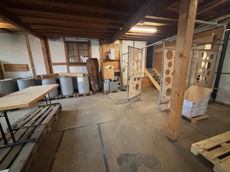 a basement, a long table on pallet, a few stacked barrels, a few stacked crates, some scattered wooden beams, and metal bars with windows