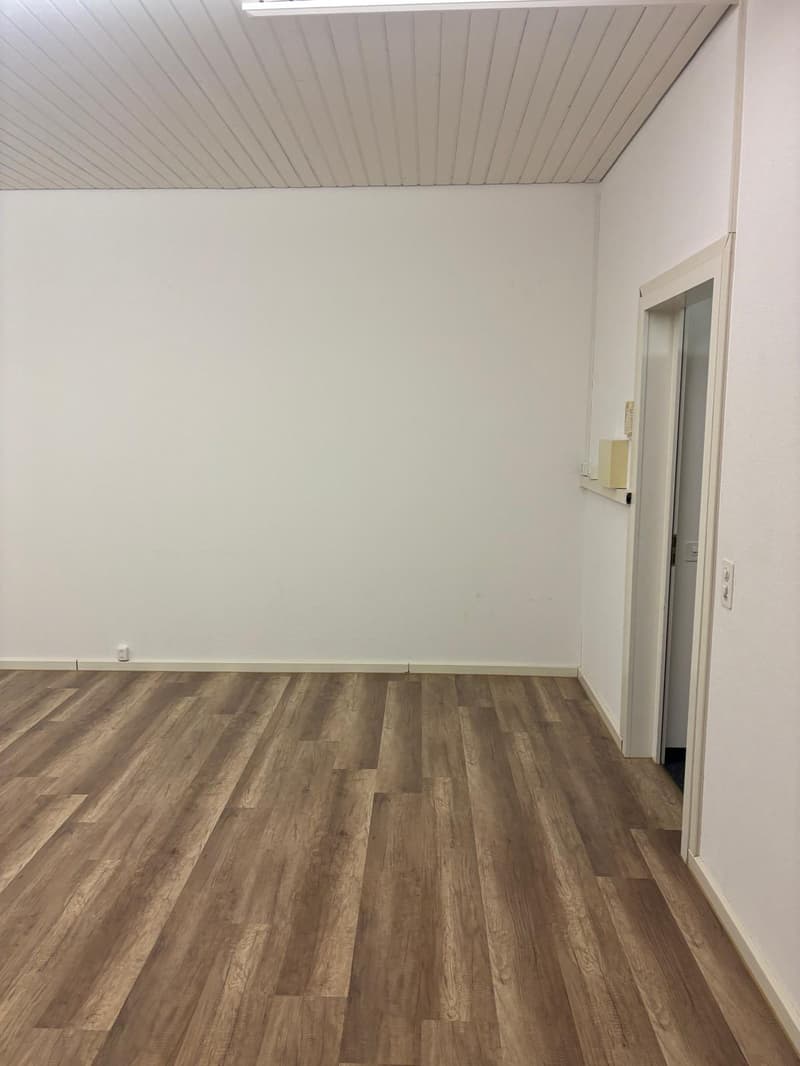The image shows a room with white walls, a paneled ceiling, and a hardwood-style laminate floor. The room appears to be empty, with a doorway visible on the right side of the image.