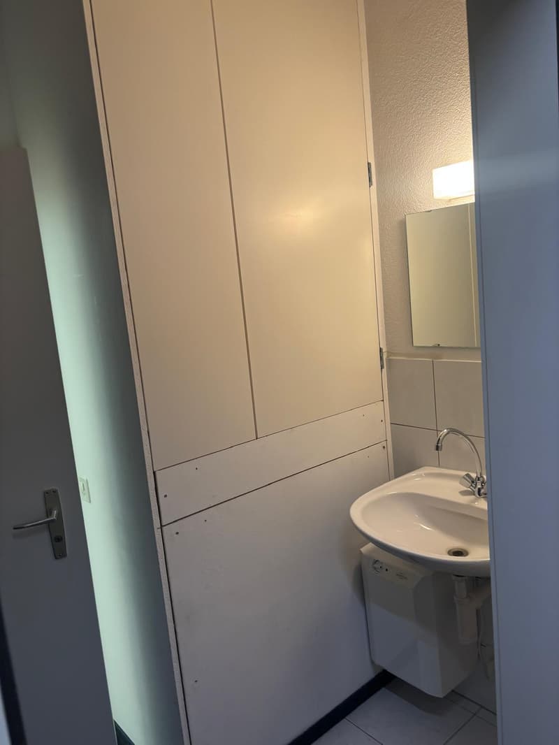 The image shows a small bathroom with a white sink and a white cabinet or storage unit on the wall. The walls appear to be a light color, and there is a door visible in the background.