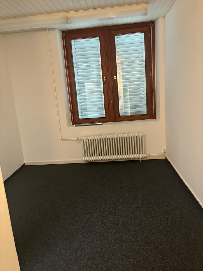 The image shows an empty room with a wooden window frame, white walls, and a black carpeted floor. There is a radiator visible in the room.