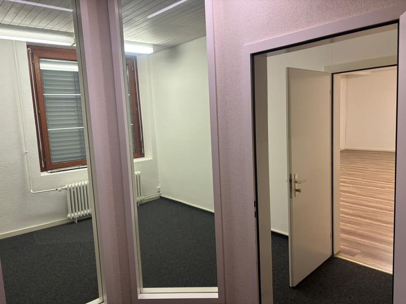 The image shows an office space with a window, a door, and a hallway leading to another room. The walls are painted in a light color, and the floor is covered with dark carpeting. The space appears to be well-lit and organized.