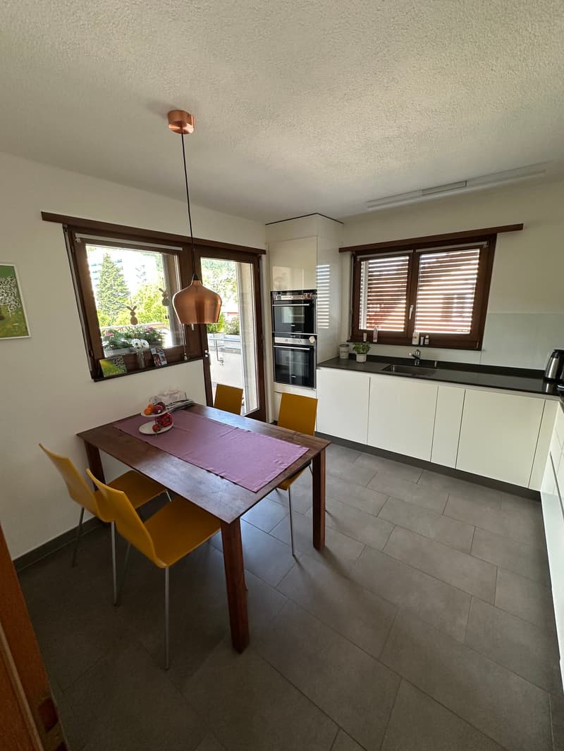 The image shows a modern, open-plan kitchen and dining area. The kitchen features white cabinets, black countertops, and built-in appliances including a dishwasher. The dining area has a wooden table with purple chairs. Large windows provide natural light