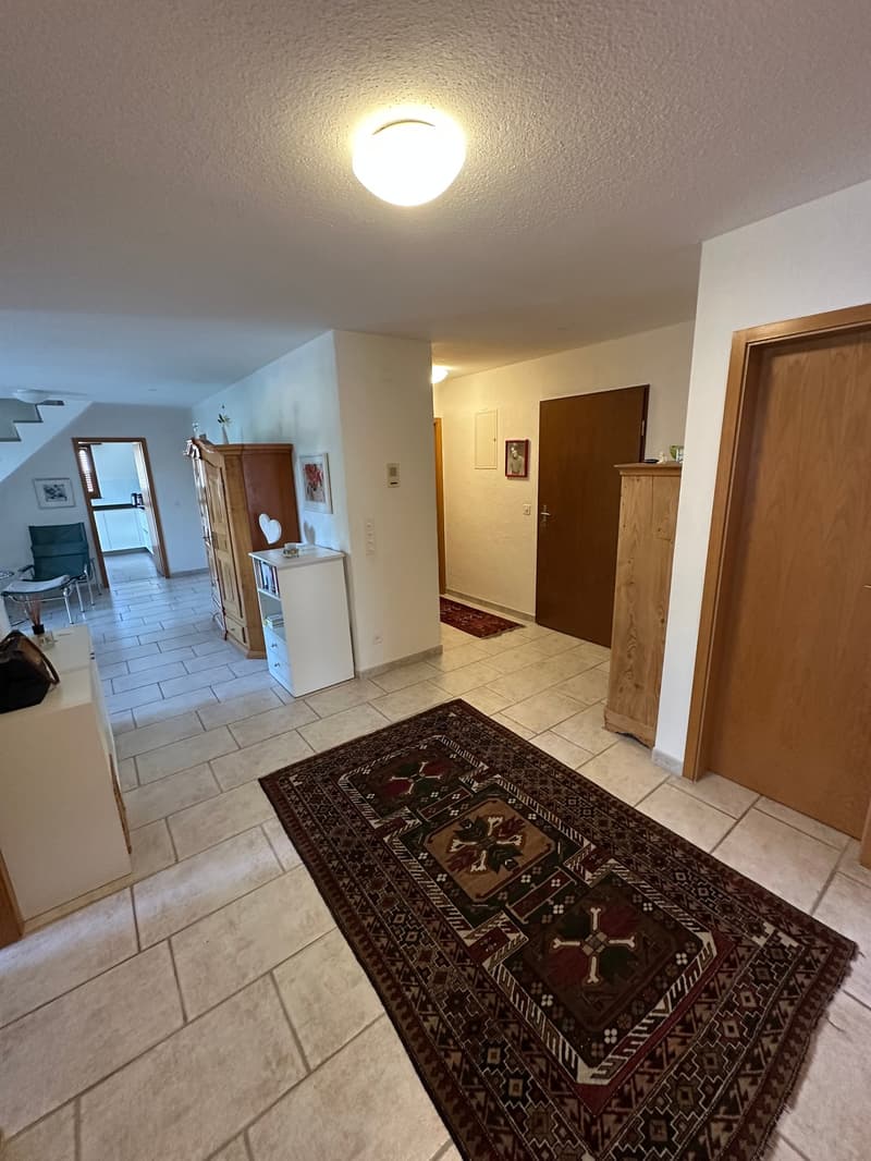 The image shows the entryway of a home. The floor is tiled with light-colored tiles, and there is a large, patterned area rug in the center. The walls are painted a neutral color, and there is a wooden cabinet or armoire visible in the background. The ent