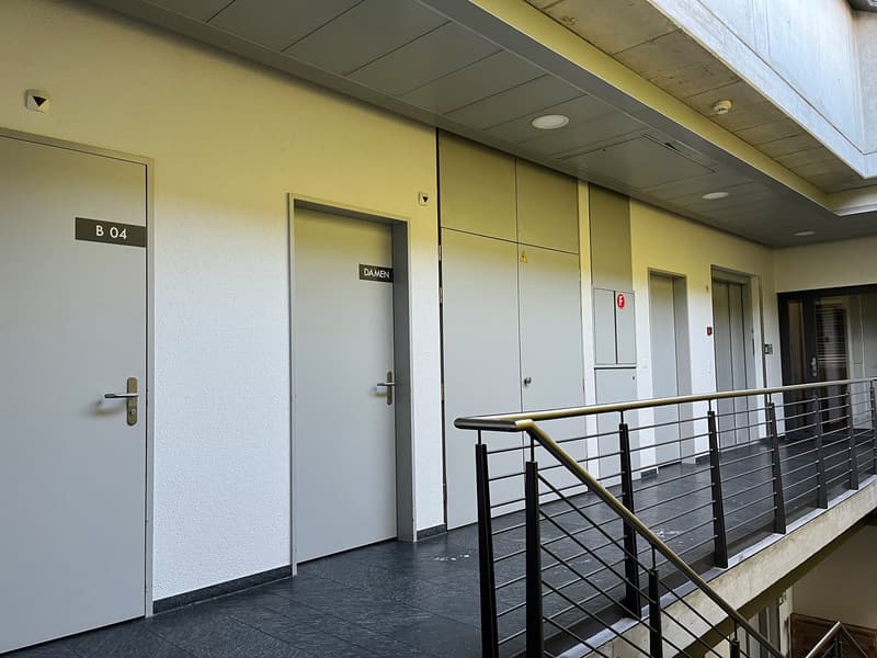 The image shows a hallway or corridor in a building. The walls are painted in a light yellow color, and the ceiling has recessed lighting. There are several doors along the hallway, one of which is labeled 'B 04' and another is labeled 'DAMEN'. The floor