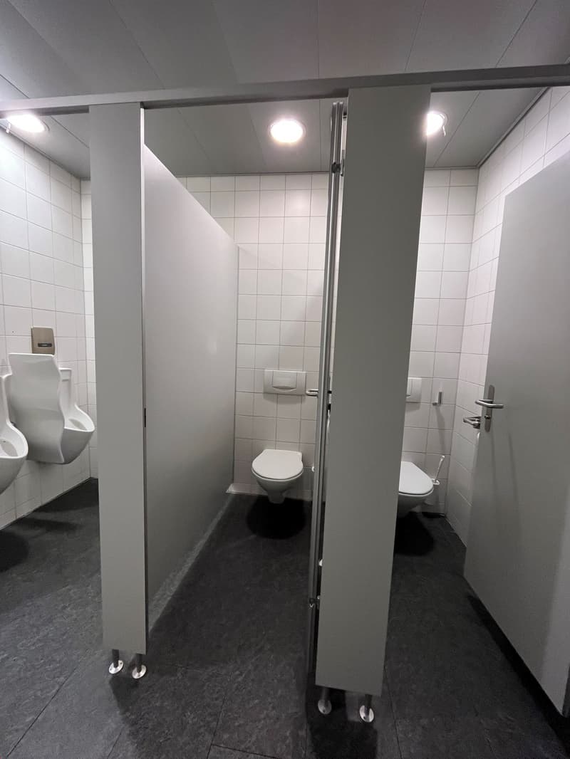 The image shows a bathroom with white tiled walls, a toilet, and a stall with a glass door. The bathroom has a modern, minimalist design with recessed lighting and a dark tile floor.