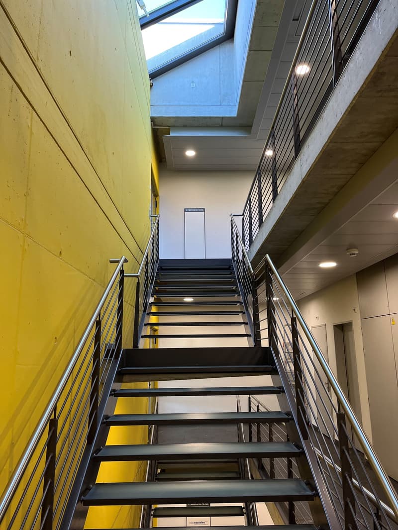 The image shows a staircase with metal railings and steps. The walls are painted in a bright yellow color, and there is a large window at the top of the staircase, allowing natural light to enter the space. The staircase appears to be located in a commerc