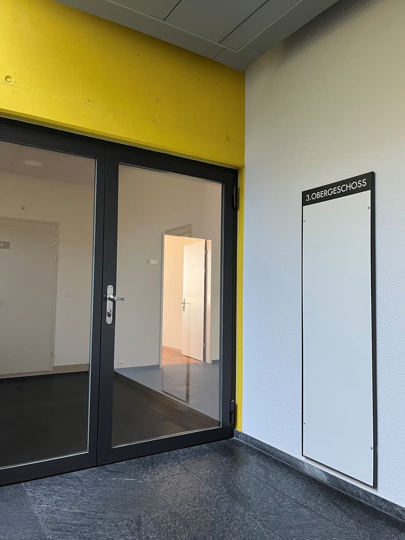 The image shows a hallway with a yellow wall, black framed glass doors, and a sign indicating the 3rd floor. The floor appears to be made of a dark gray material.