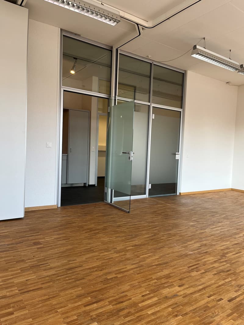 Spacious office with hardwood flooring, glass partition walls, and a glass door. The room has track lighting and appears to be a modern, well-designed workspace.