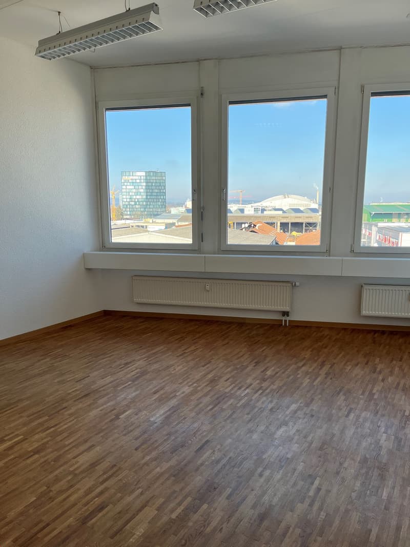 Large open office space with hardwood flooring, white walls, and large windows providing a view of the city skyline outside.
